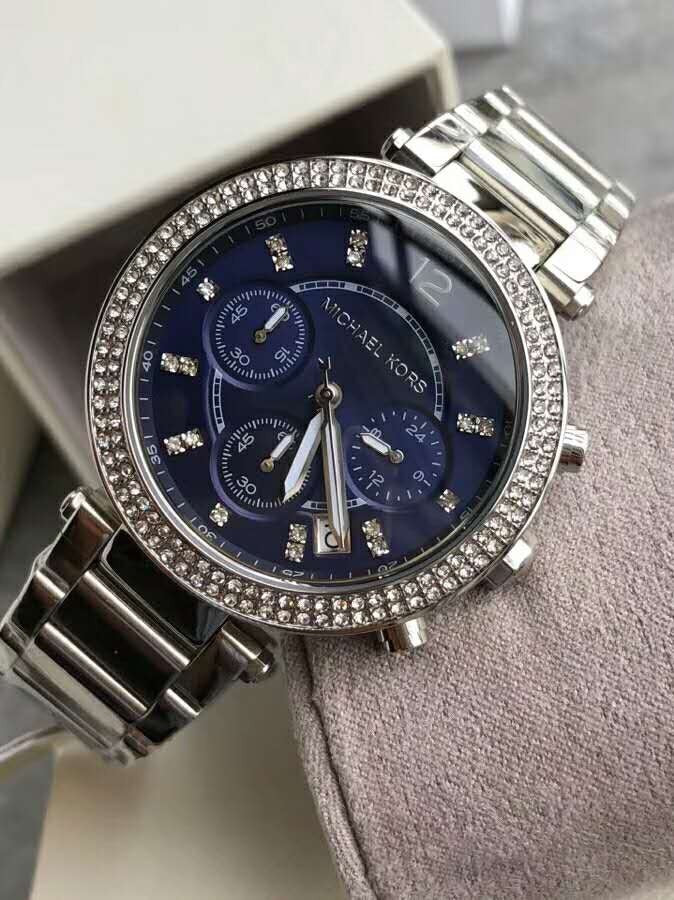 Close-up display shot of the Michael Kors MK6117 watch resting on a light grey velvet cushion, highlighting the reflective blue dial.