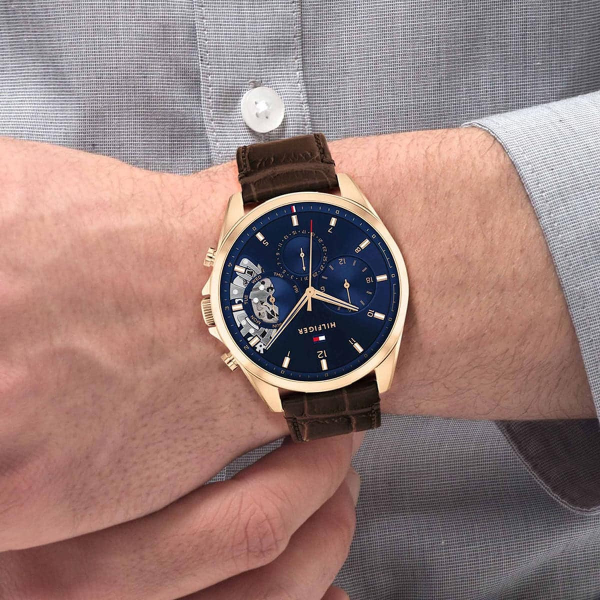 Man wearing the Tommy Hilfiger 1710453 Baker watch on his wrist, styled with a grey dress shirt to demonstrate the 44mm case size and fit.